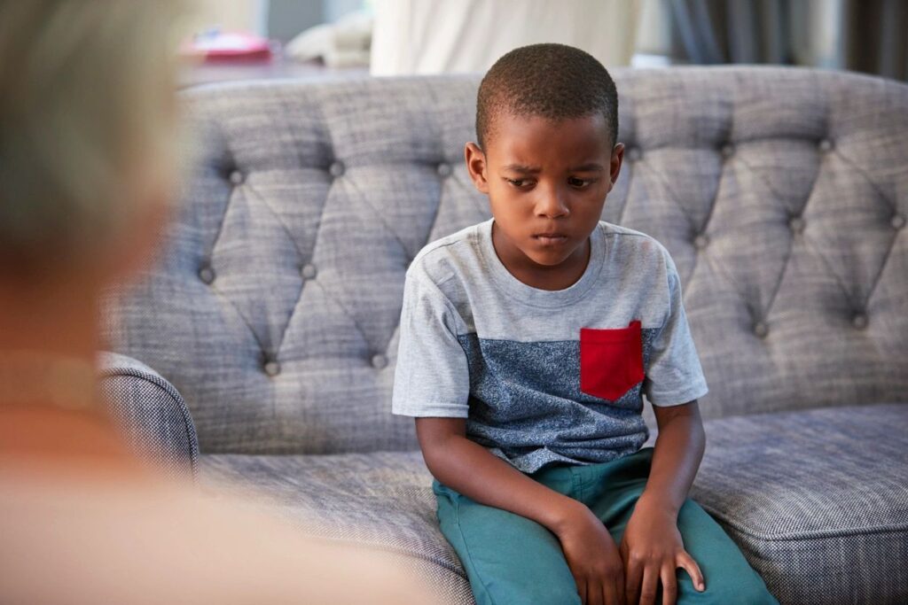 a little boy is sitting on a couch. His is looking down and away from the adult in the background and appears sad.