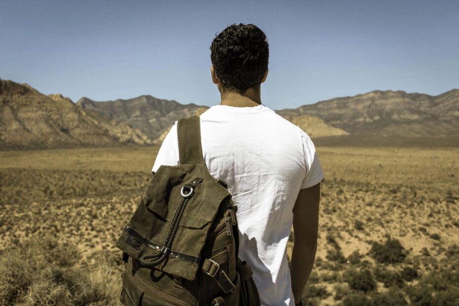 man standing with his back to camera facing a mountain range. Man has a backpack on his left shoulder